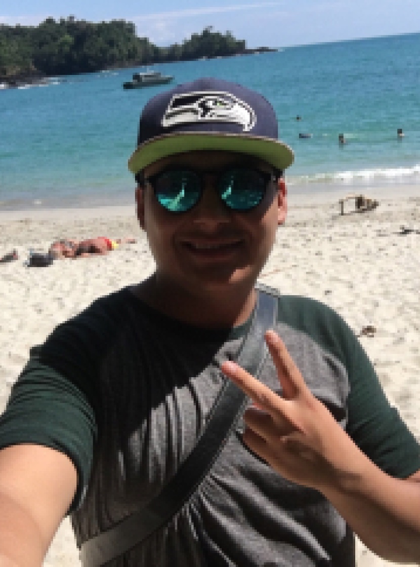 Yamil taking a selfie on a beach, wearing a cap and sunglasses and making a peace sign.